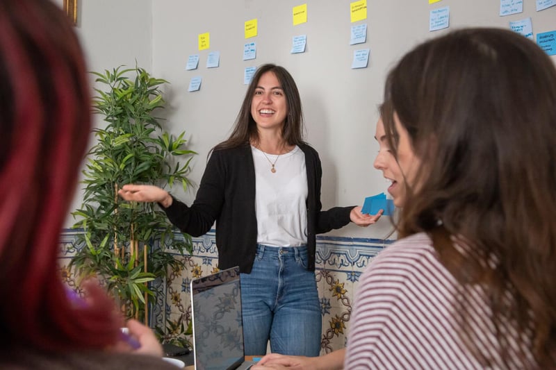 Image shows a smiling young woman standing in front of a white board with post its while other women look at a laptop