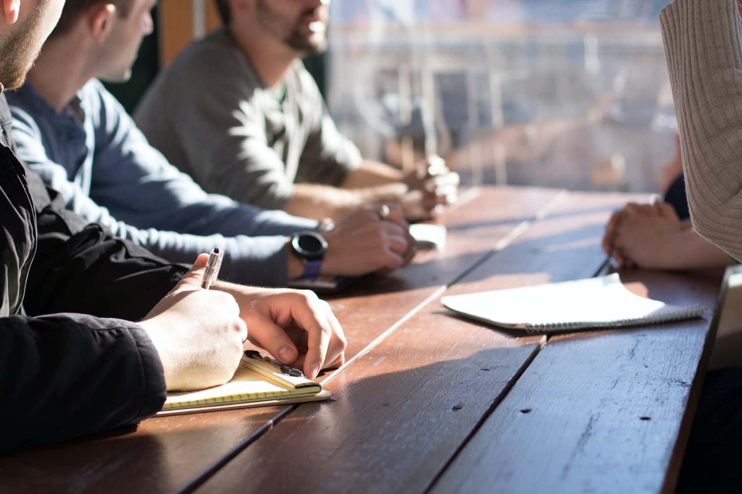 Image shows three people sitting at a wooden table taking notes while someone stands facing them
